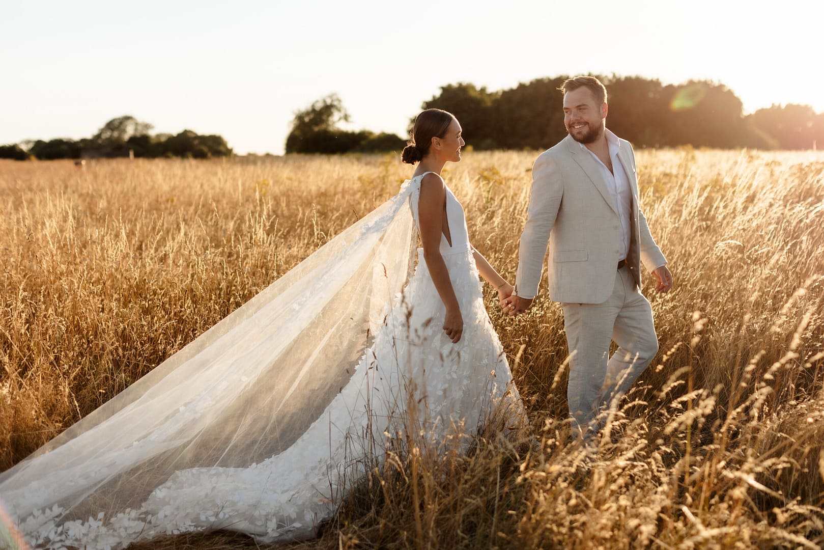 Bride and groom walking through field at sunset, photographed by London wedding photographer